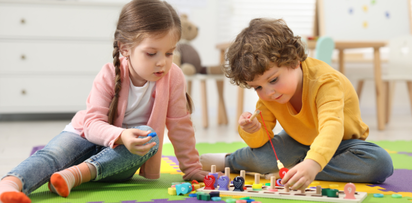 Two young children playing with a wooden toy on the floor.]