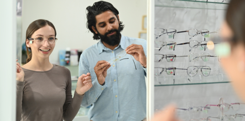 A happy young woman trying on spectacles at an opticians while a male holds another pair of glasses
